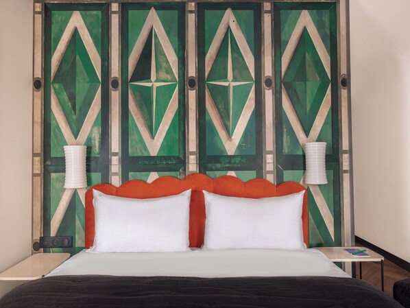 A bed with white and orange bedding, a nightstand with a lamp, and a geometric wall design. - Chachacha Hôtel (Nantes)