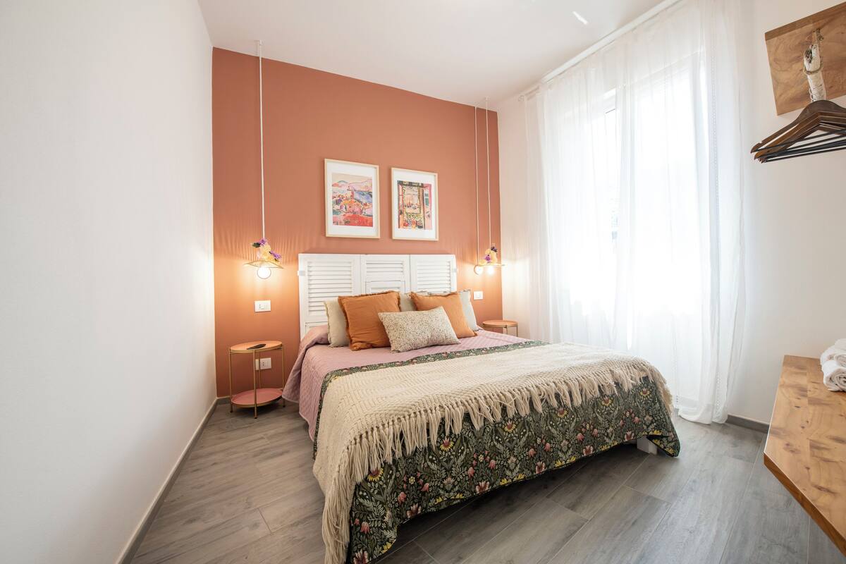 Une chambre à coucher avec un lit, deux tables de chevet, un luminaire suspendu et une fenêtre avec des rideaux.