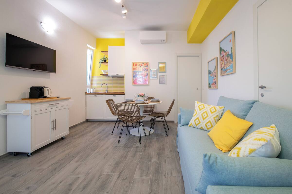 Un salon moderne avec un canapé bleu, un coin repas avec des chaises en osier et une cuisine équipée d’une télévision et d’une machine à café.