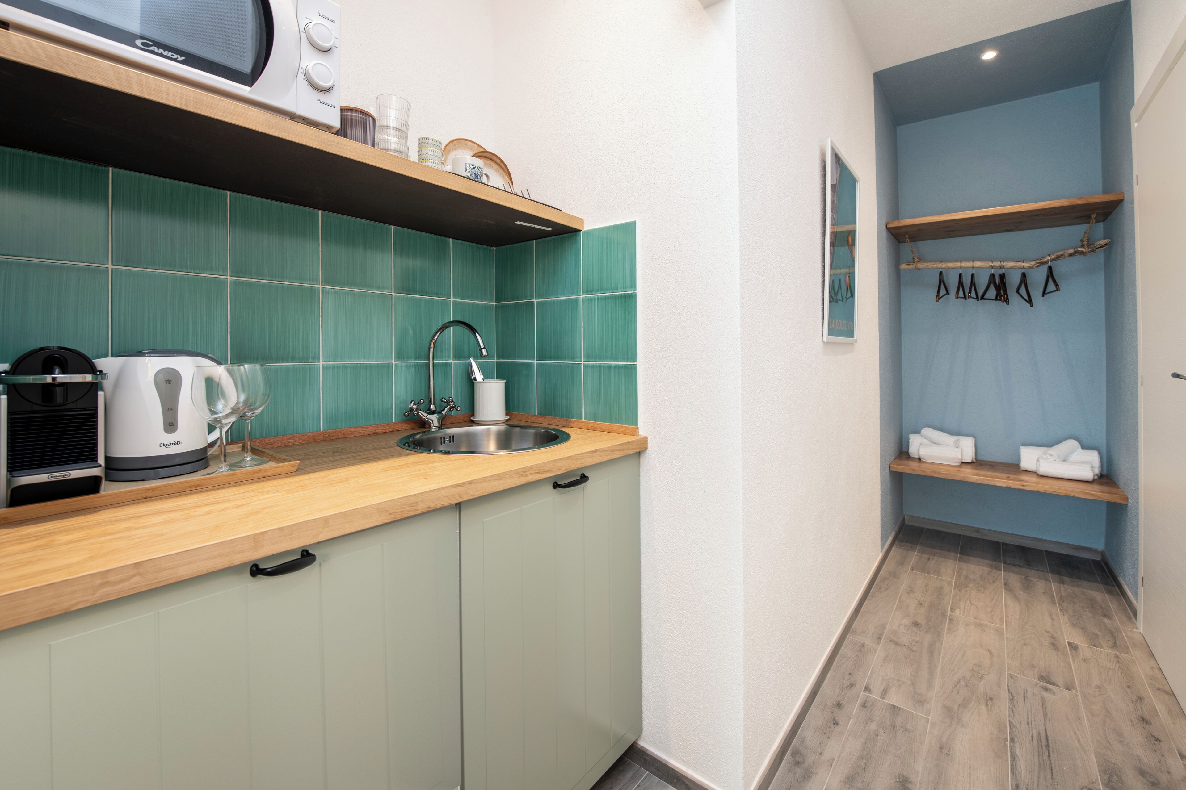 Een kleine keuken met een spoelbak, waterkoker en broodrooster. Daarboven zijn houten planken bij de spoelbak en een handdoekhouder aan de muur.