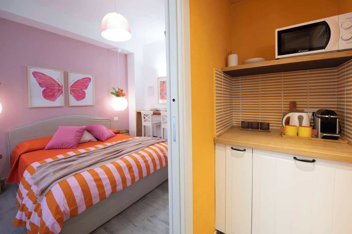 Une chambre à coucher avec un lit, une table de chevet et une commode. Il y a une fenêtre avec des rideaux, un mur orné d’un cadre, et une porte qui donne sur une autre pièce.