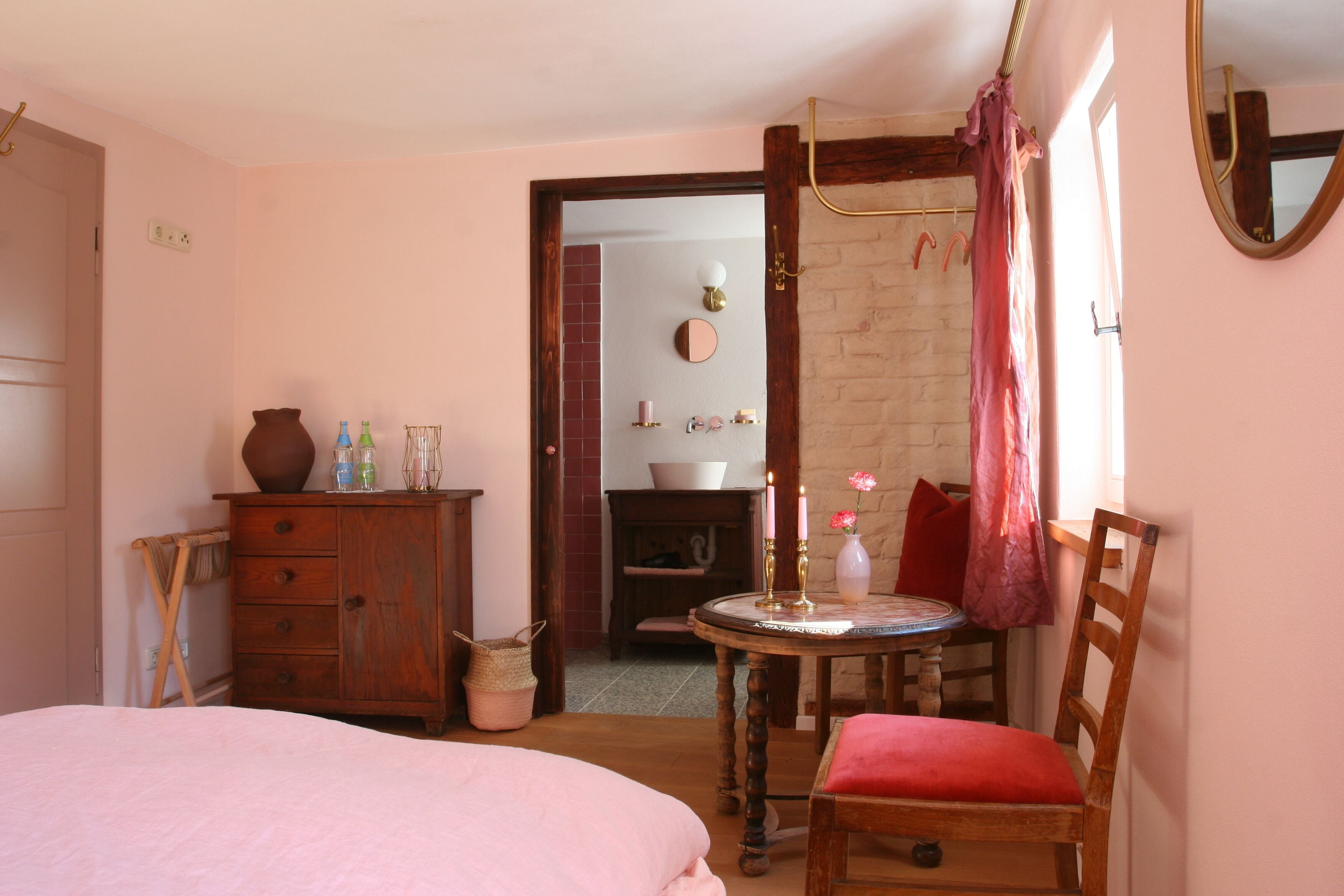 A bedroom with a bed, a wooden dresser, a small table with a chair, a mirror, and a fireplace in the background.