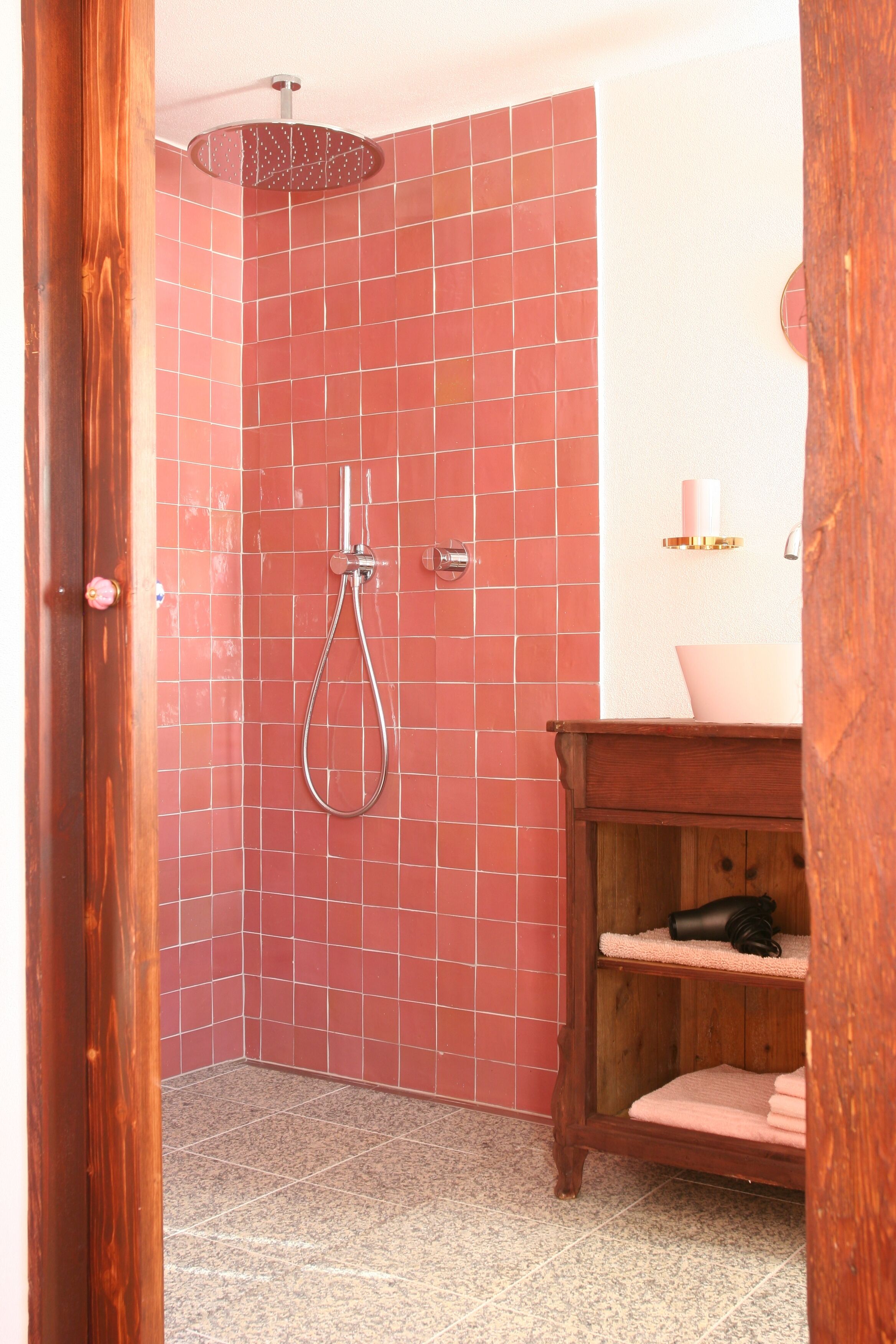 A bathroom with red tiled walls, a wooden vanity, a sink, and a shower with a handheld showerhead.
