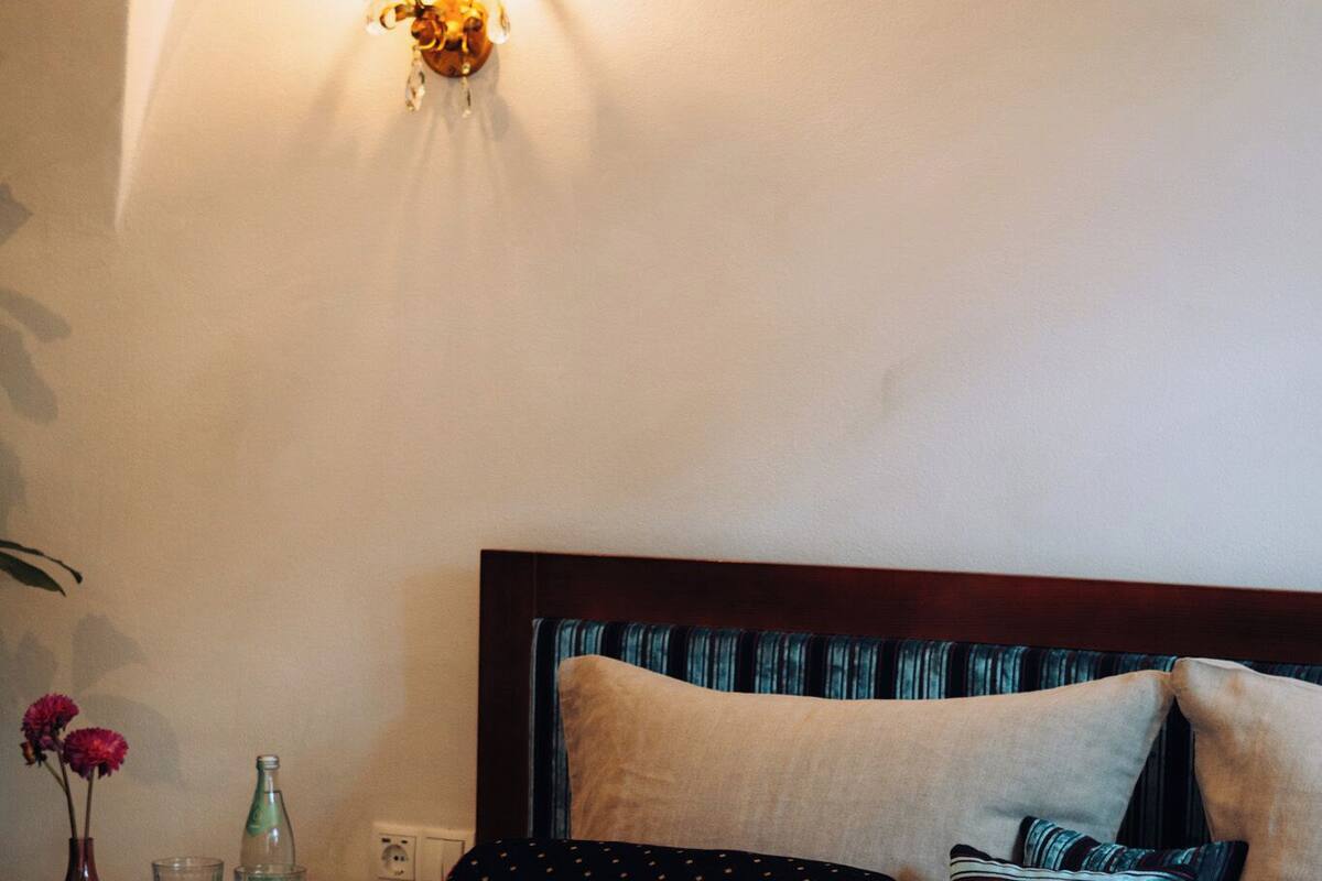 A neatly made bed with a white duvet, a wooden headboard, and several pillows. There is a vase with flowers and a bottle on the nightstand beside the bed.
