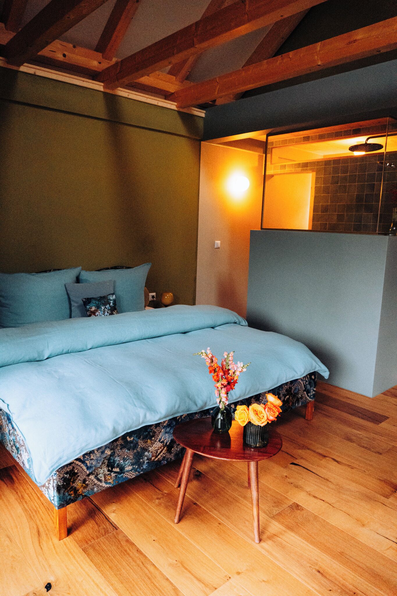 Une chambre avec un grand lit, une petite table ronde ornée d’un vase de fleurs, un plancher en bois et une poutre apparente au plafond.