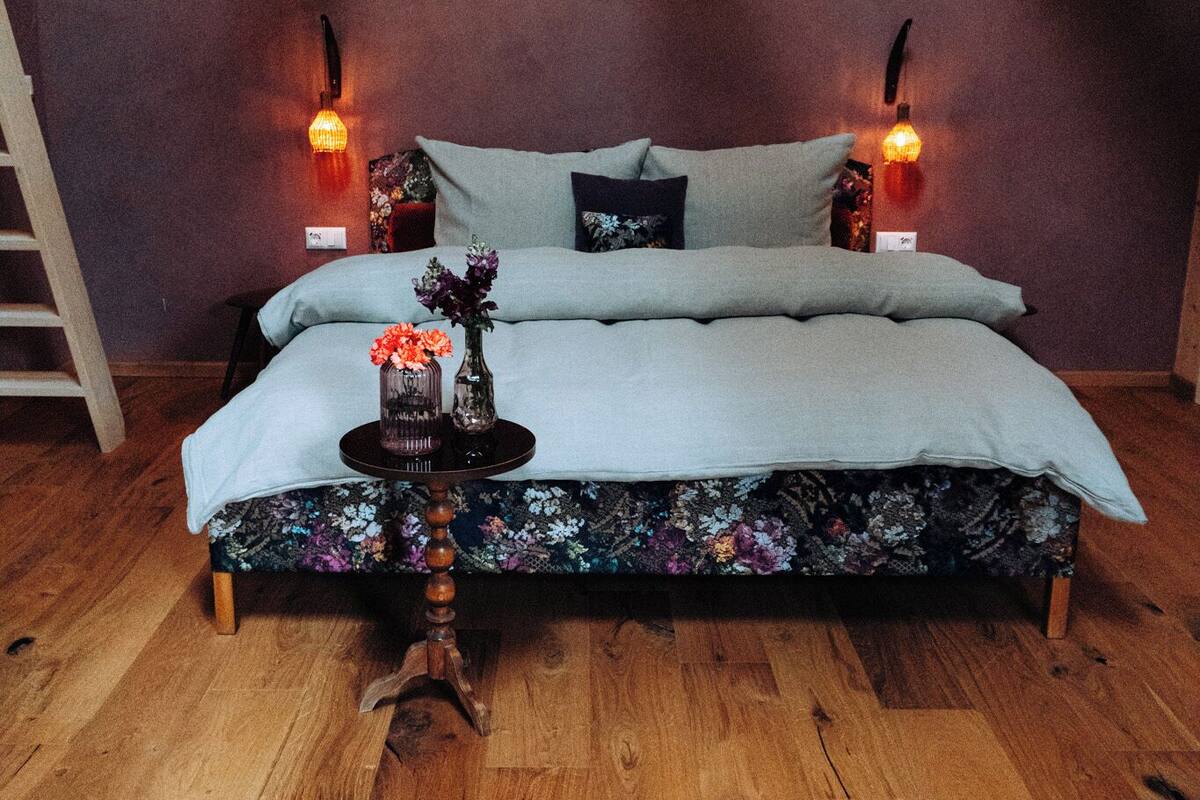 A bedroom with a wooden floor, a bed with floral bedding, two bedside lamps, and a small table with a vase.