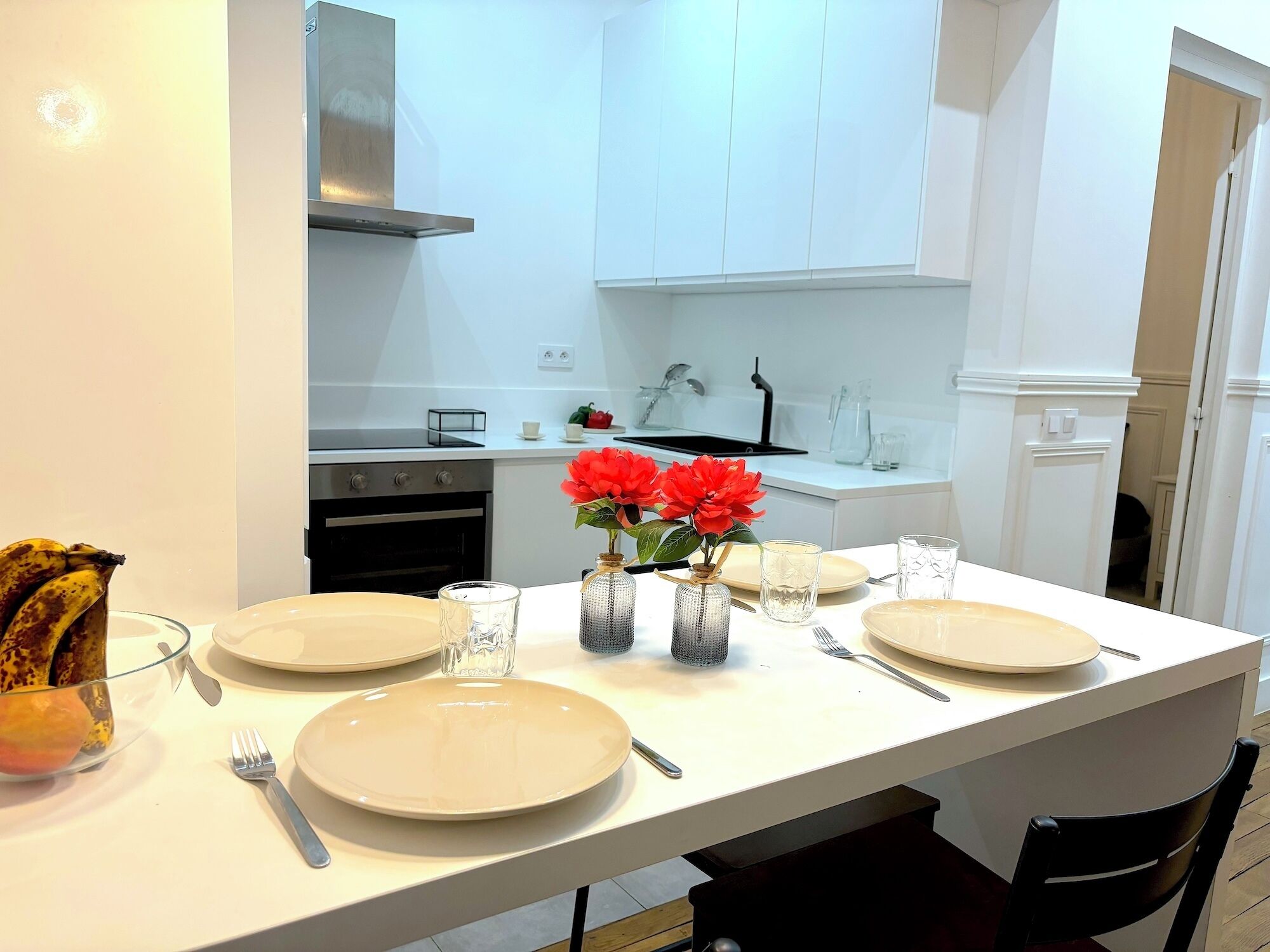Una cocina moderna con armarios blancos, un extractor de acero inoxidable y una mesa de comedor con platos, vasos y un jarrón con flores rojas.