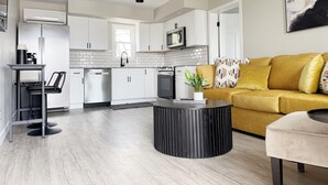 Eine moderne Küche mit weißen Schränken, einer Mikrowelle und einer Kaffeemaschine. Ein gelbes Sofa mit gemusterten Kissen und ein schwarzer Couchtisch sind ebenfalls vorhanden.