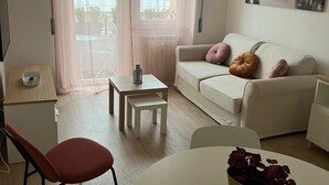 En moderne stue med sofa, spisebord og stol. Der er et vindue med let gardin, et lille bord med et lys, og et billede på væggen.
