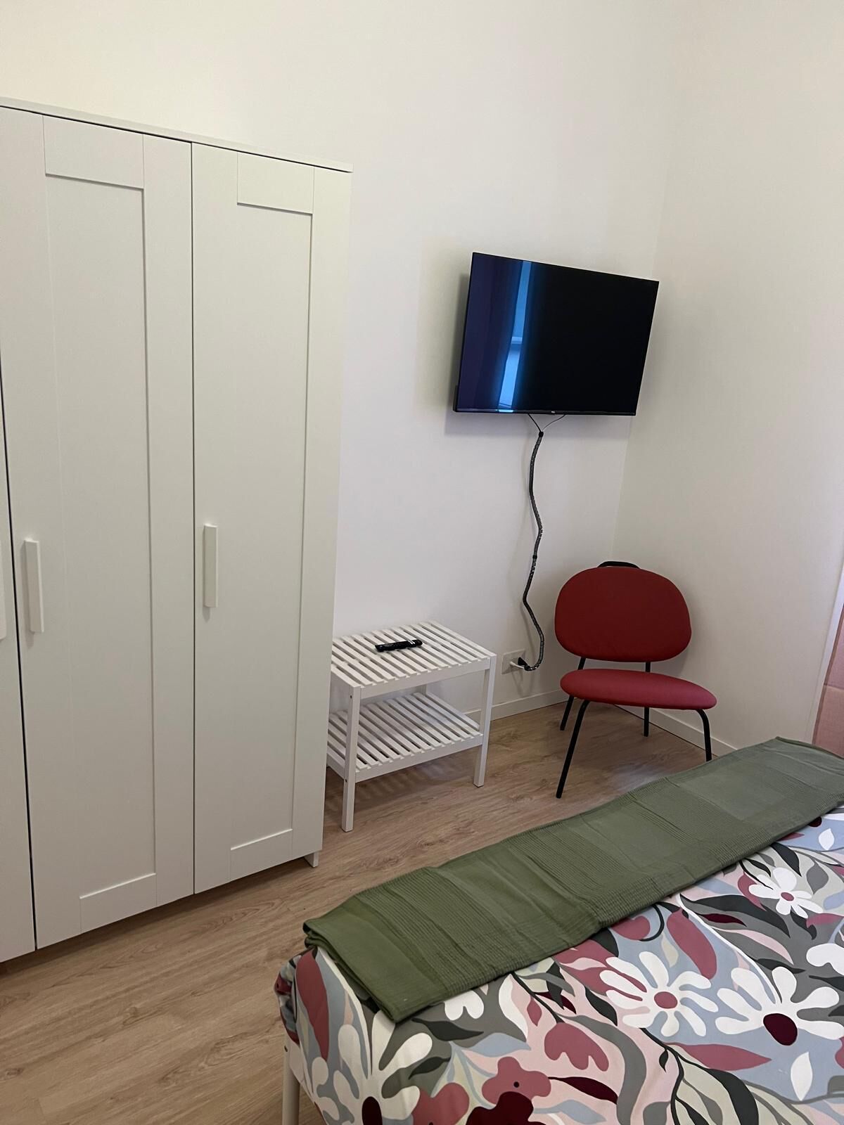 Une chambre avec une armoire, un téléviseur fixé au mur, un fauteuil rouge et un lit recouvert d’une couvre-lit à motifs floraux.