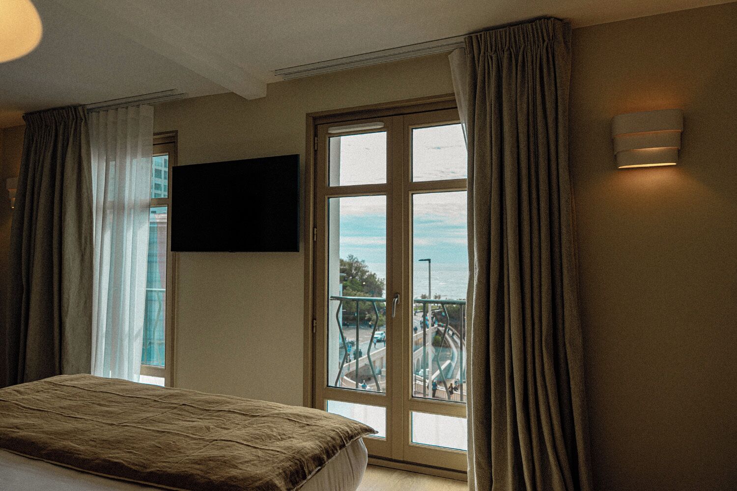 A hotel room with a bed, a TV mounted on the wall, a balcony with a view of the city, and a window with curtains.