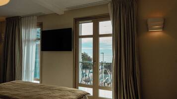 A hotel room with a bed, a TV mounted on the wall, a balcony with a view of the city, and a window with curtains.