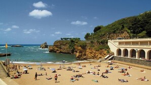 A beach with people sunbathing, a rocky coastline, and a building with arches. - Roc Seven Biarritz (Biarritz)