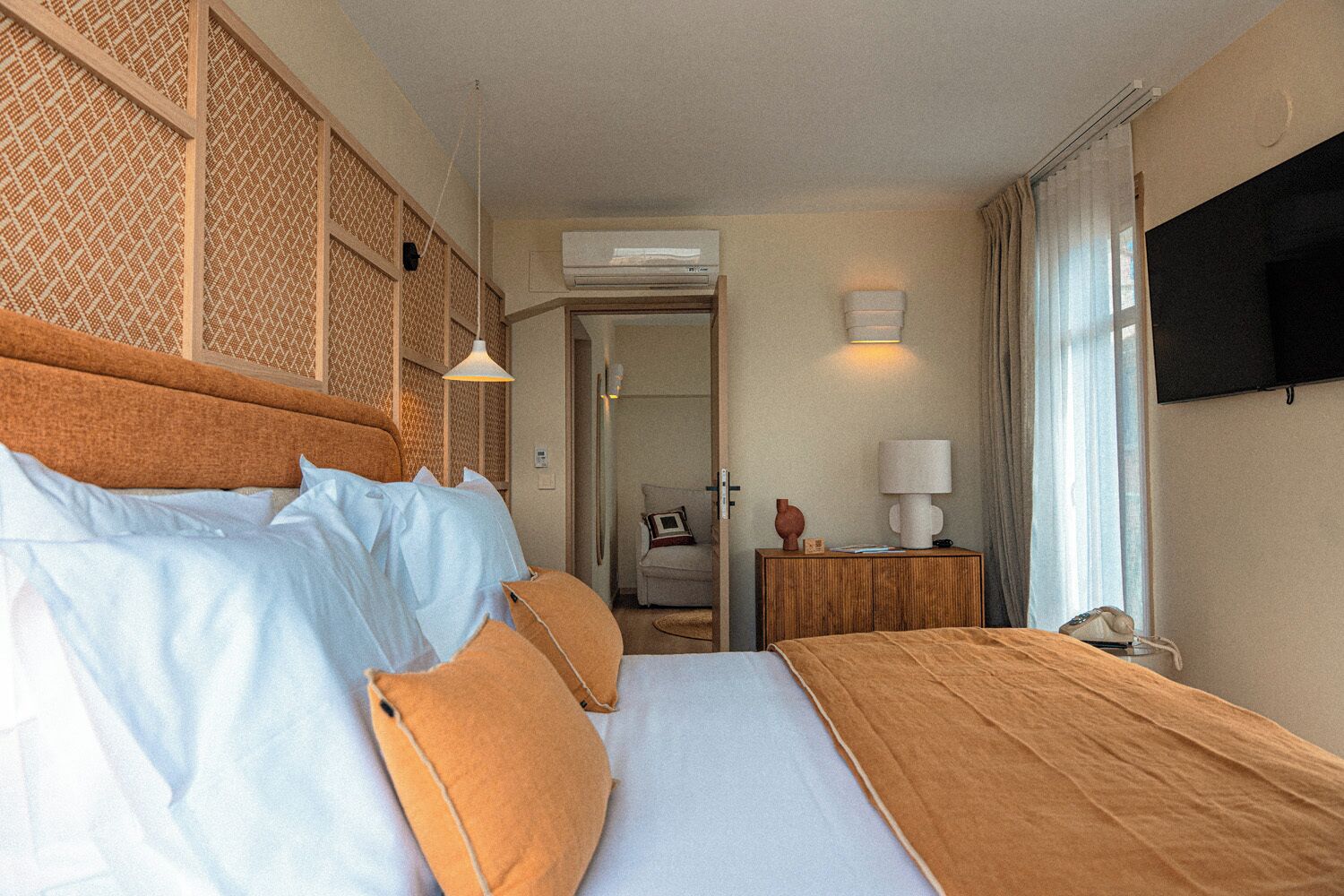 A hotel room with a large bed, a wooden nightstand, a wall-mounted TV, and a door leading to another room.
