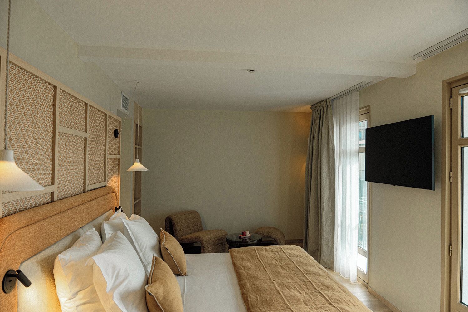 A hotel room with a bed, bedside table, lamp, chair, TV, and a window with curtains.