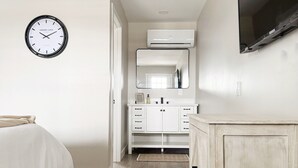 A bedroom with a bed, a wall-mounted clock, a TV, and a bathroom with a mirror and cabinets. - RiverStone Retreat D (Newburgh)