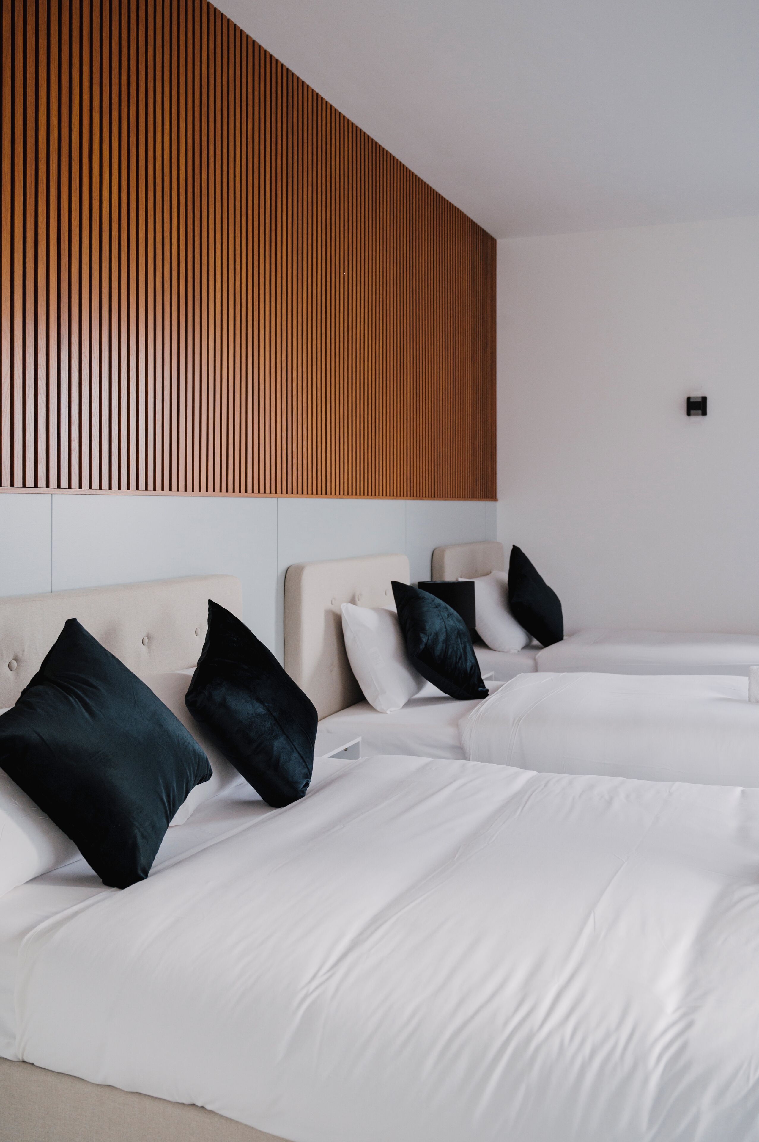 A modern hotel room with two beds, each with white bedding and black pillows. The room features a wooden slat wall design and a small black switch on the wall.