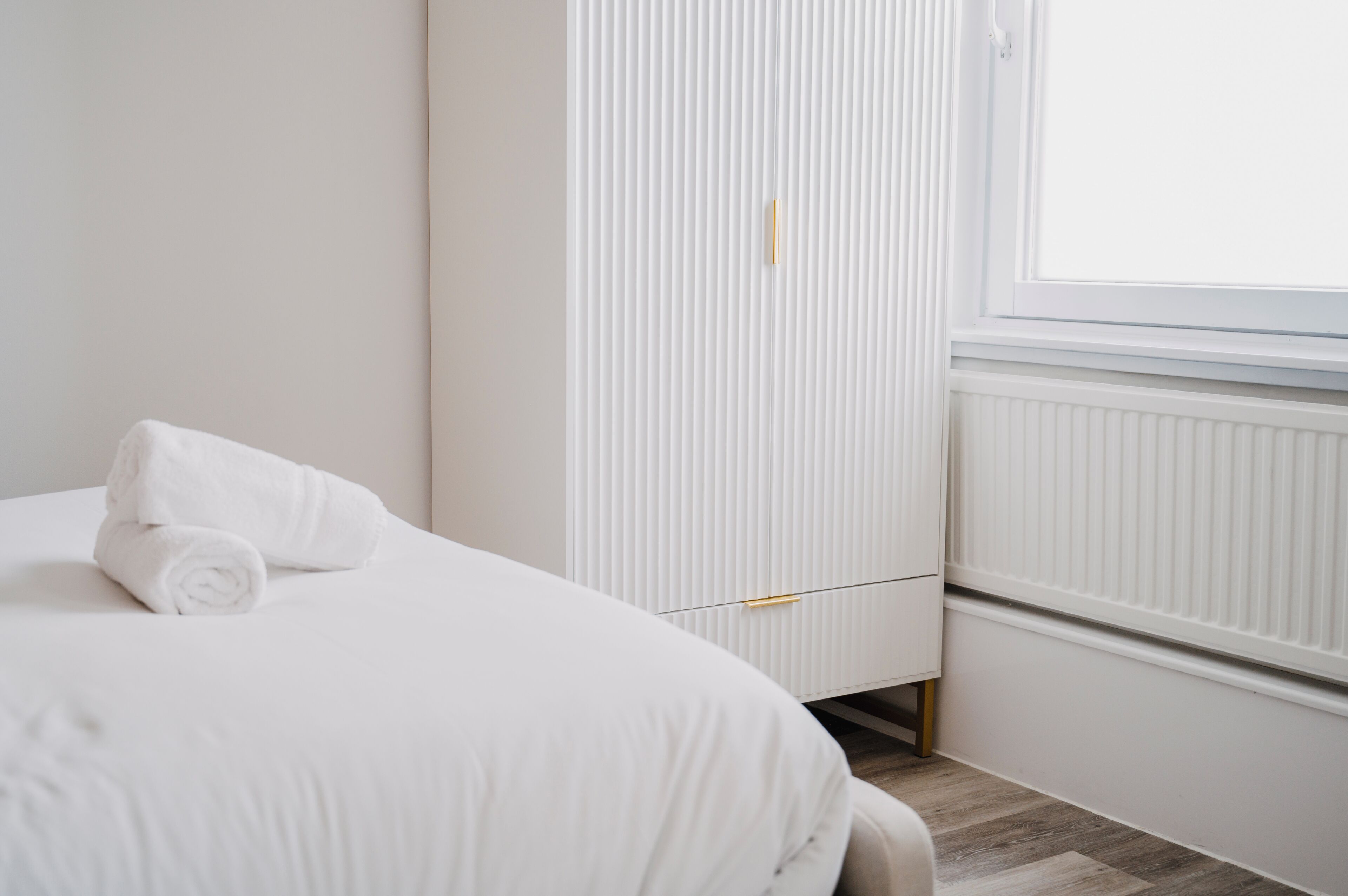 A neatly made bed with a rolled white towel, a white wardrobe with a gold handle, and a window with blinds.