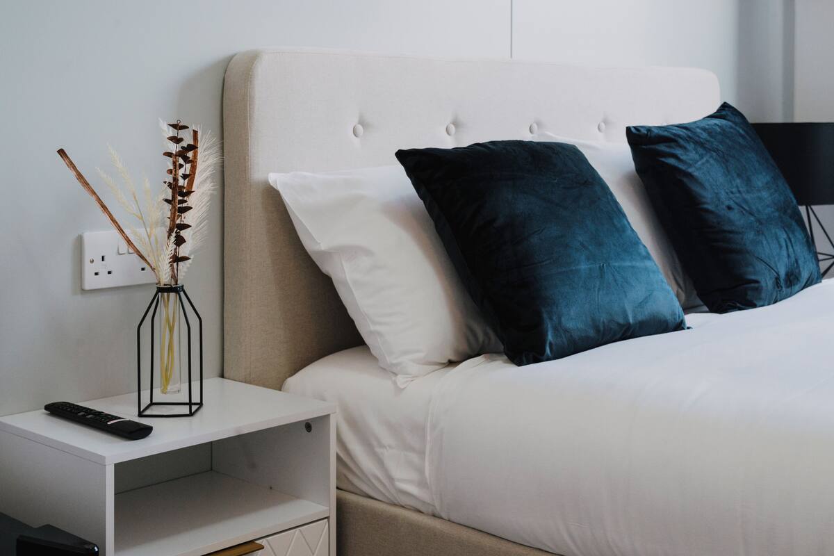 A neatly made bed with white and dark blue pillows, a white nightstand with a remote control, and a vase with dried plants.