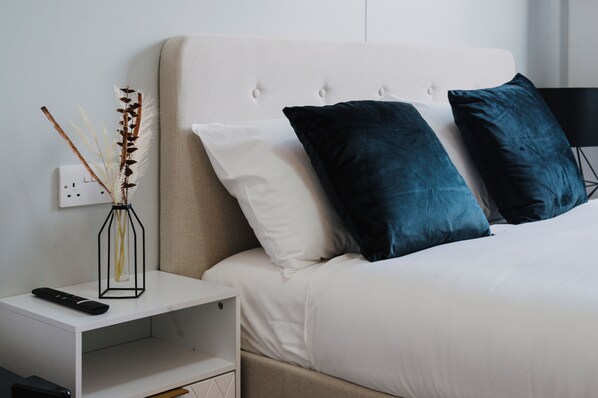 Una cama bien hecha con almohadas blancas y azul oscuro, una mesita de noche blanca con un control remoto y un florero con plantas secas.