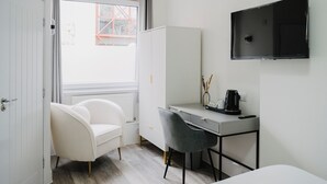 A compact hotel room with a white wardrobe, a desk with a chair, a bed, a TV mounted on the wall, and a window with a view of a building outside. - WNS Hotel (London)