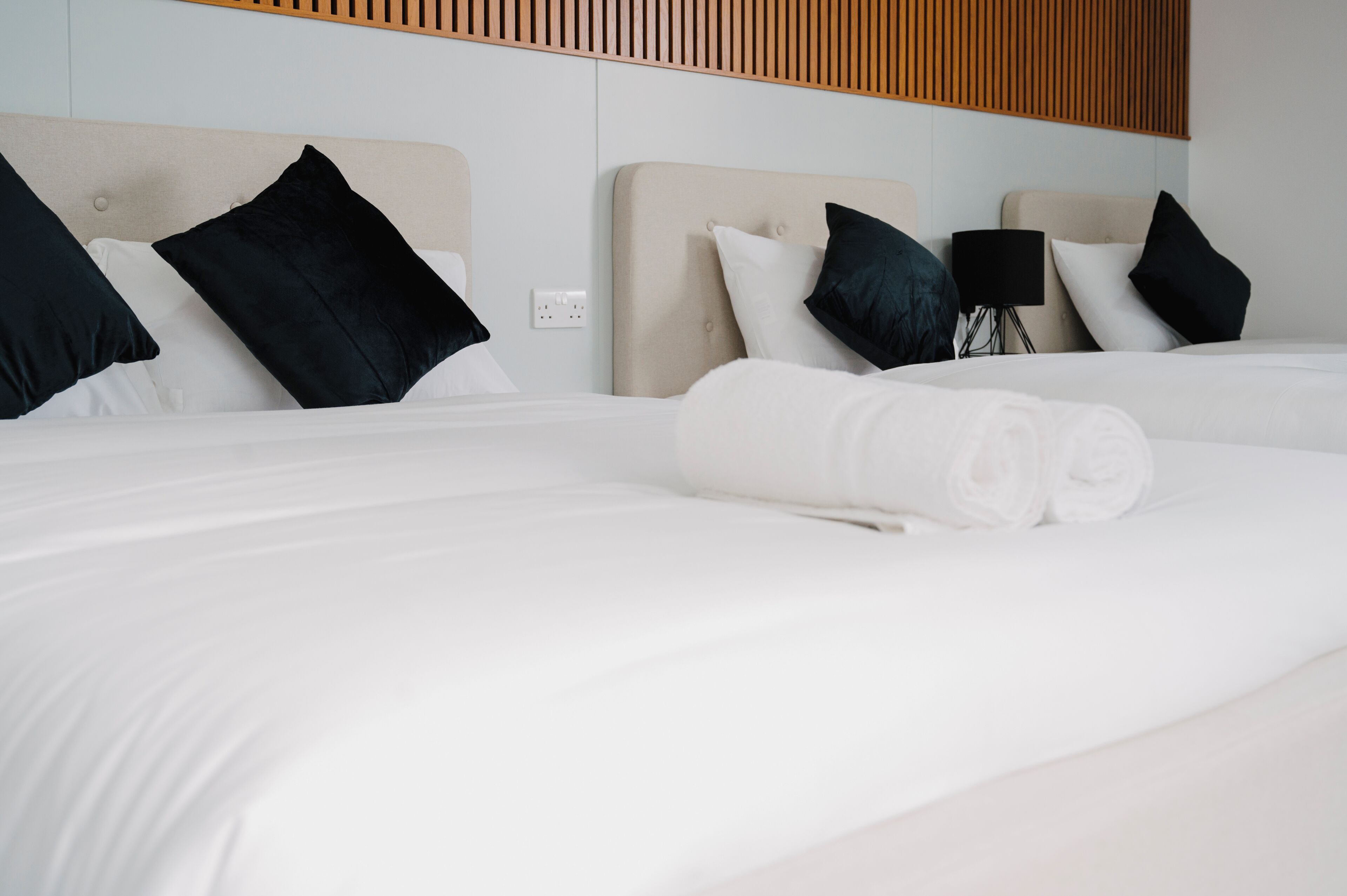 A neatly made bed with white linens and rolled towels, a headboard with a light-colored fabric, and a black floor lamp beside the bed.