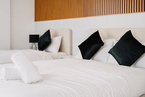 Habitación de hotel con dos camas, cada una con ropa de cama blanca y almohadas negras. Hay una pared de listones de madera sobre las camas y una mesita de noche con una lámpara al lado de una cama.