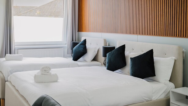 A hotel room with two beds, a wooden slat wall, a window with curtains, and a bedside table with a lamp. - WNS Hotel (London)