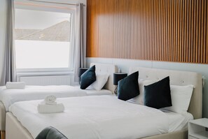 Hotellihuone, jossa on kaksi sänkyä, puusäleistä tehty seinä, ikkuna, jossa on verhot, ja yöpöytä, jossa on lamppu.