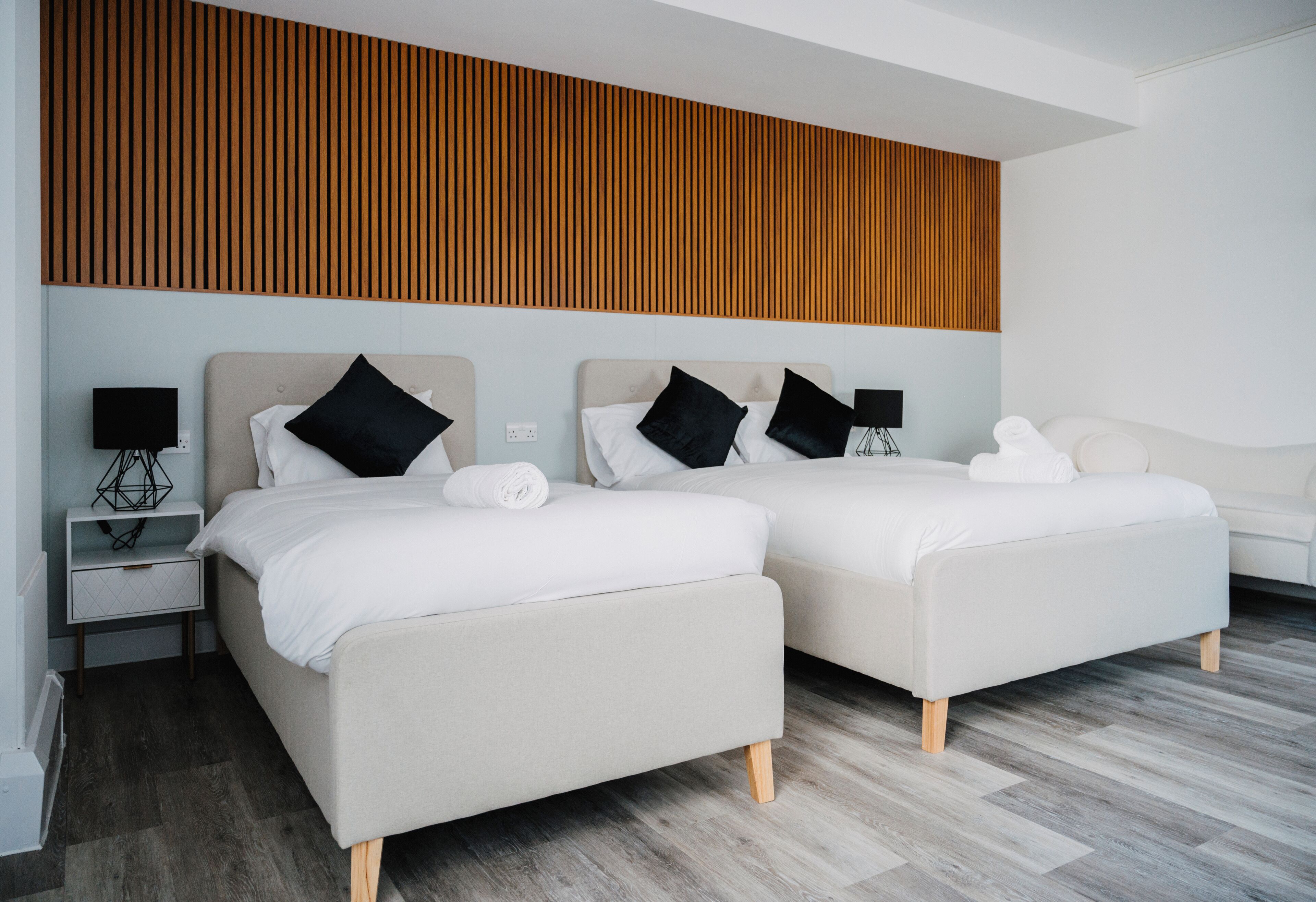 Una habitación de hotel moderna con dos camas, un panel de pared de madera, un sofá blanco y una mesita de noche con lámpara.
