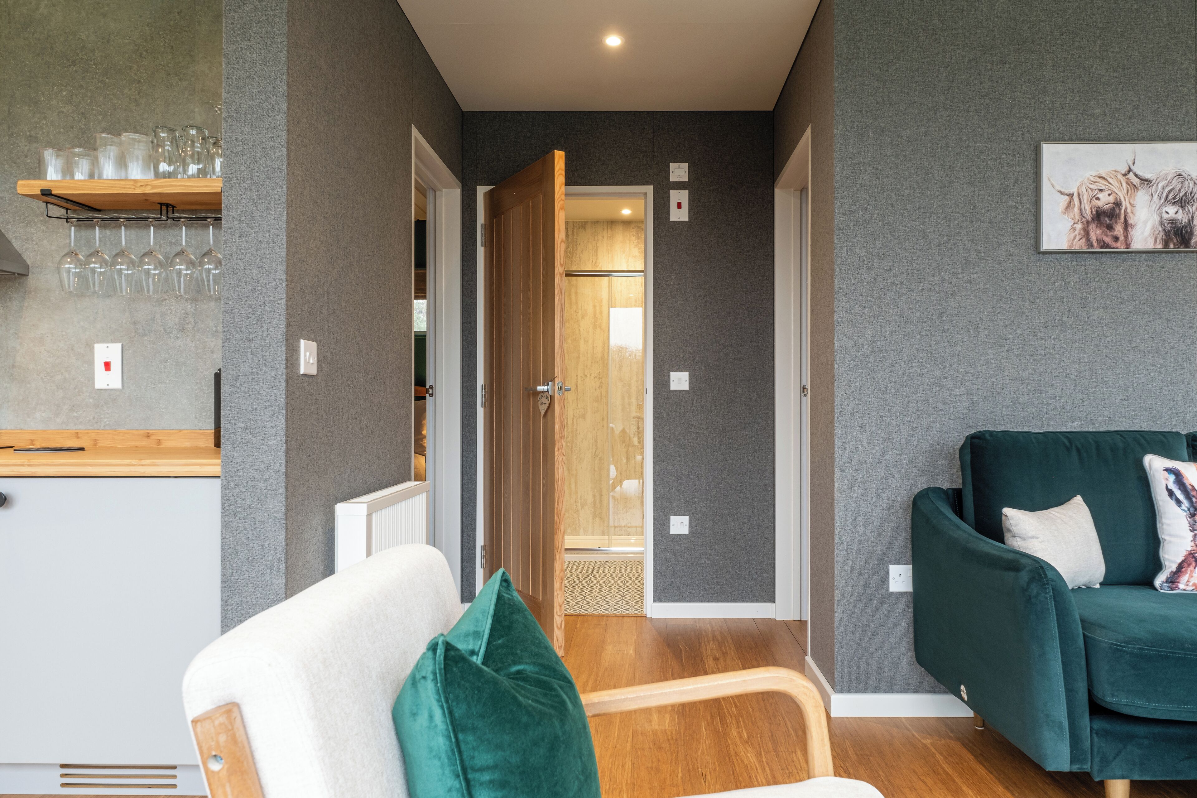 Een moderne woonkamer met een groene bank, een houten stoel en een keukenhoek met planken en glazen.