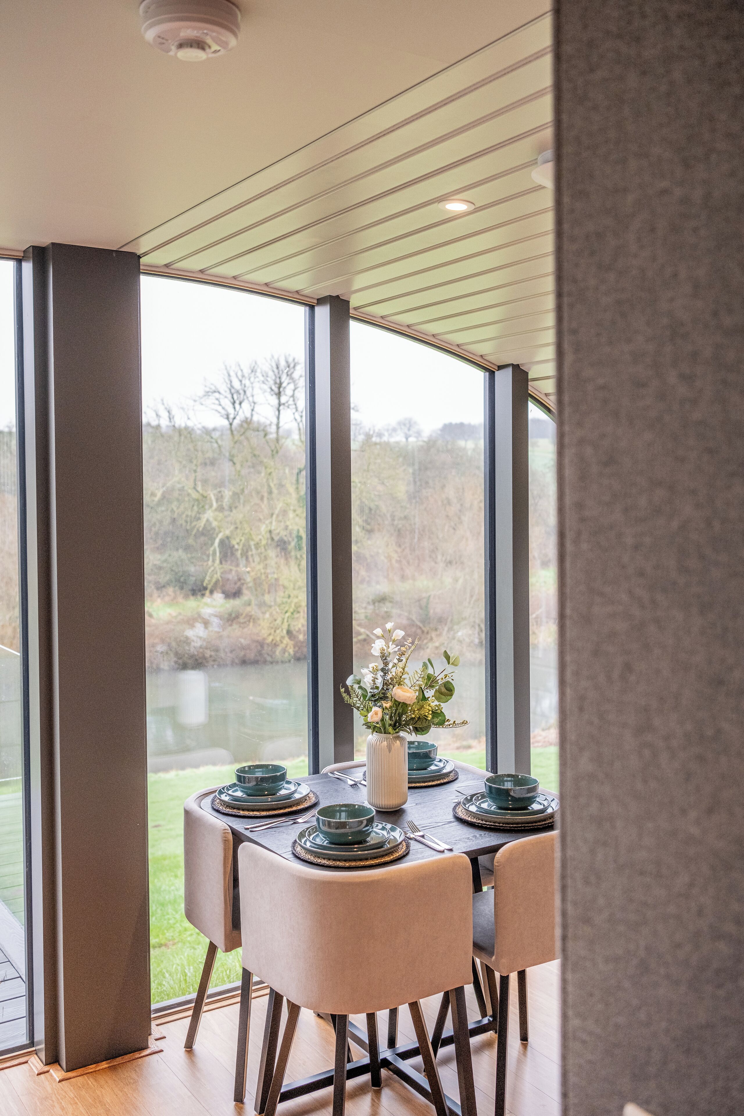 A dining area with a table set for four, large windows, and a view of a river and trees.