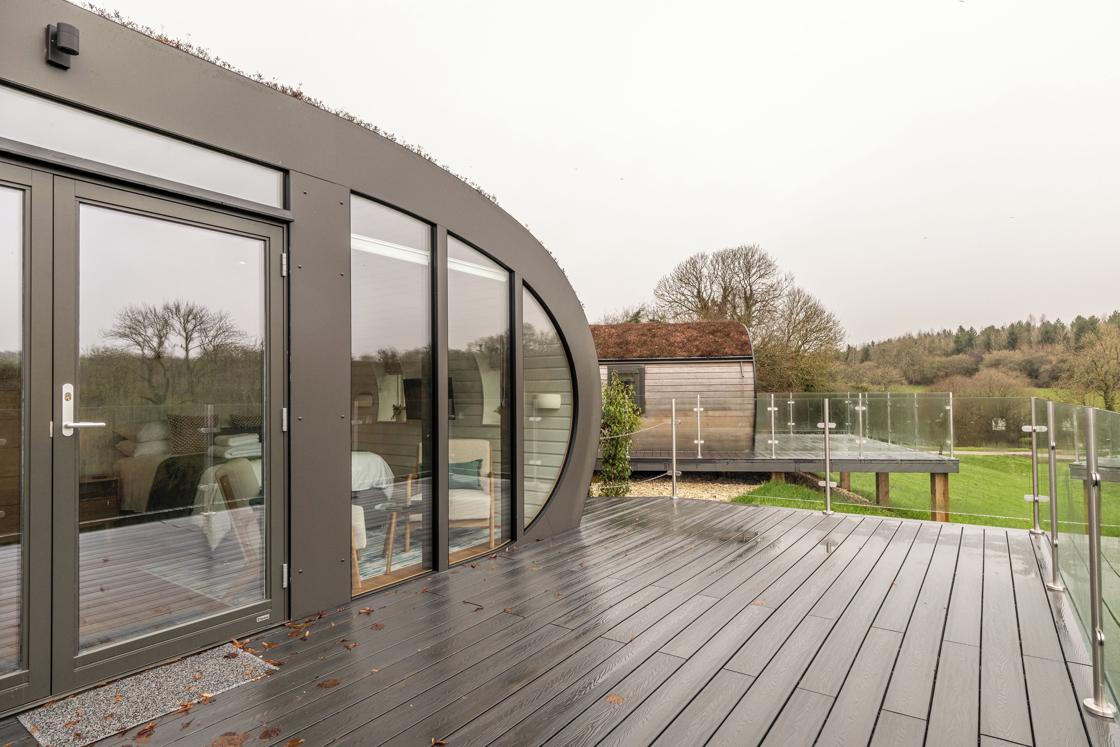 Ein modernes Haus mit einer gebogenen Glasdoor, einer Holzterrasse und einem kleinen Schuppen im Hintergrund.