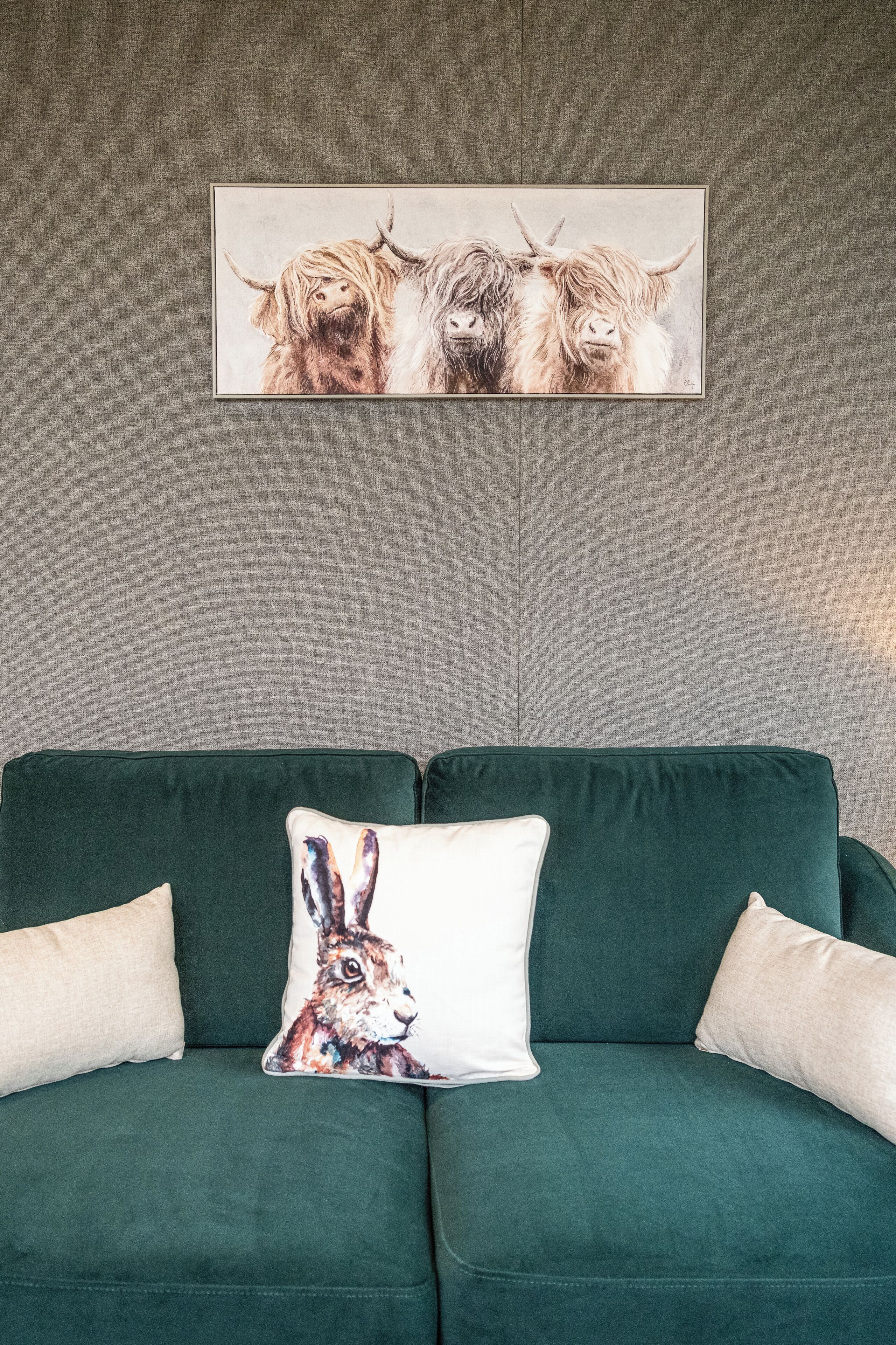 Un canapé vert avec deux coussins, dont l’un est orné d’un motif représentant un lapin. Au-dessus du canapé, on peut voir un tableau encadré représentant trois animaux.