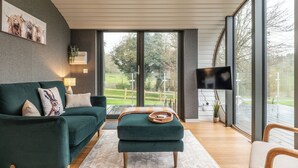 A modern living room with a green sofa, a wooden ottoman, and a TV mounted on the wall. - Sustainable 2BD Pod on Old Chalford Farm (Chipping Norton)