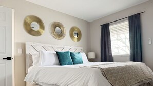 Ein ordentlich bezogenes Bett mit weißen und türkisfarbenen Kissen, ein Holz-Kopfteil und drei goldene Spiegel an der Wand.