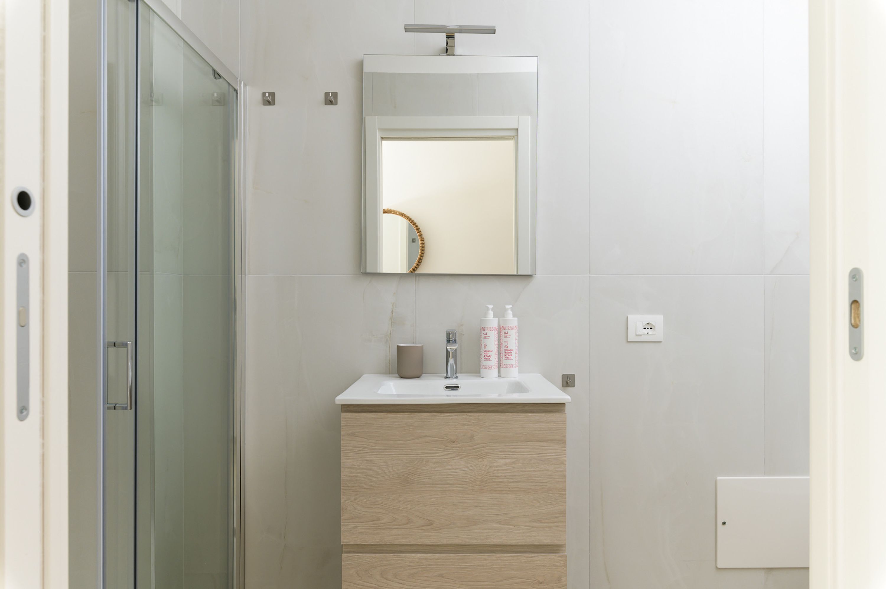 Une salle de bain moderne avec une douche à l’italienne en verre, un lavabo blanc avec un miroir au-dessus, et deux flacons de produits de toilette posés sur le lavabo.