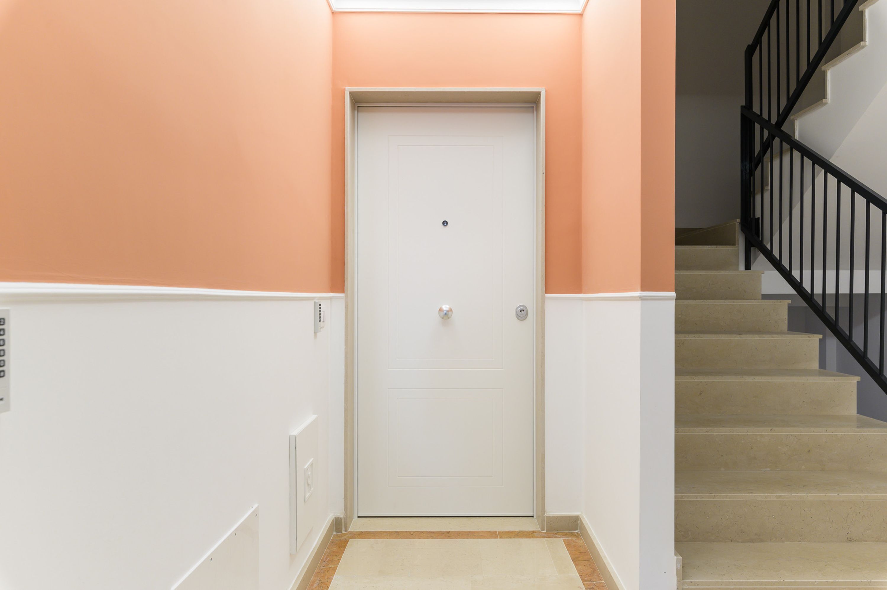 A hallway with a white door, beige carpet, and a staircase with black railings.