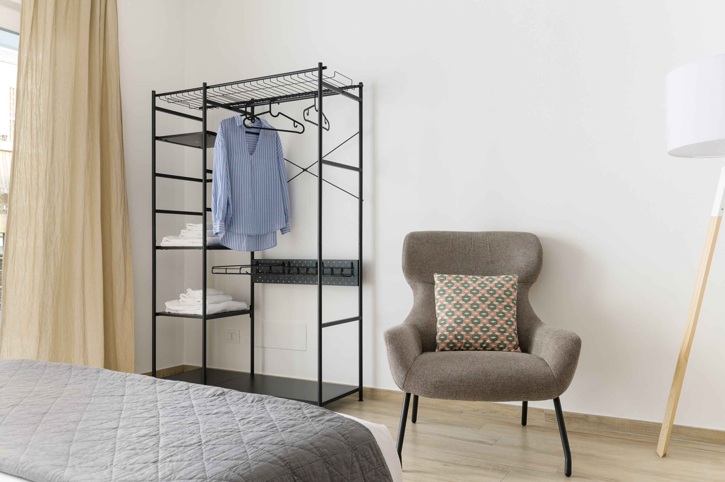 Una habitación con una cama, un sillón gris con un cojín estampado, un tendedero de metal negro con ropa y una lámpara blanca.