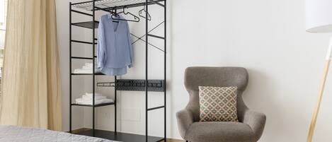 Una habitación con una cama, un sillón gris con un cojín estampado, un tendedero de metal negro con ropa y una lámpara blanca.