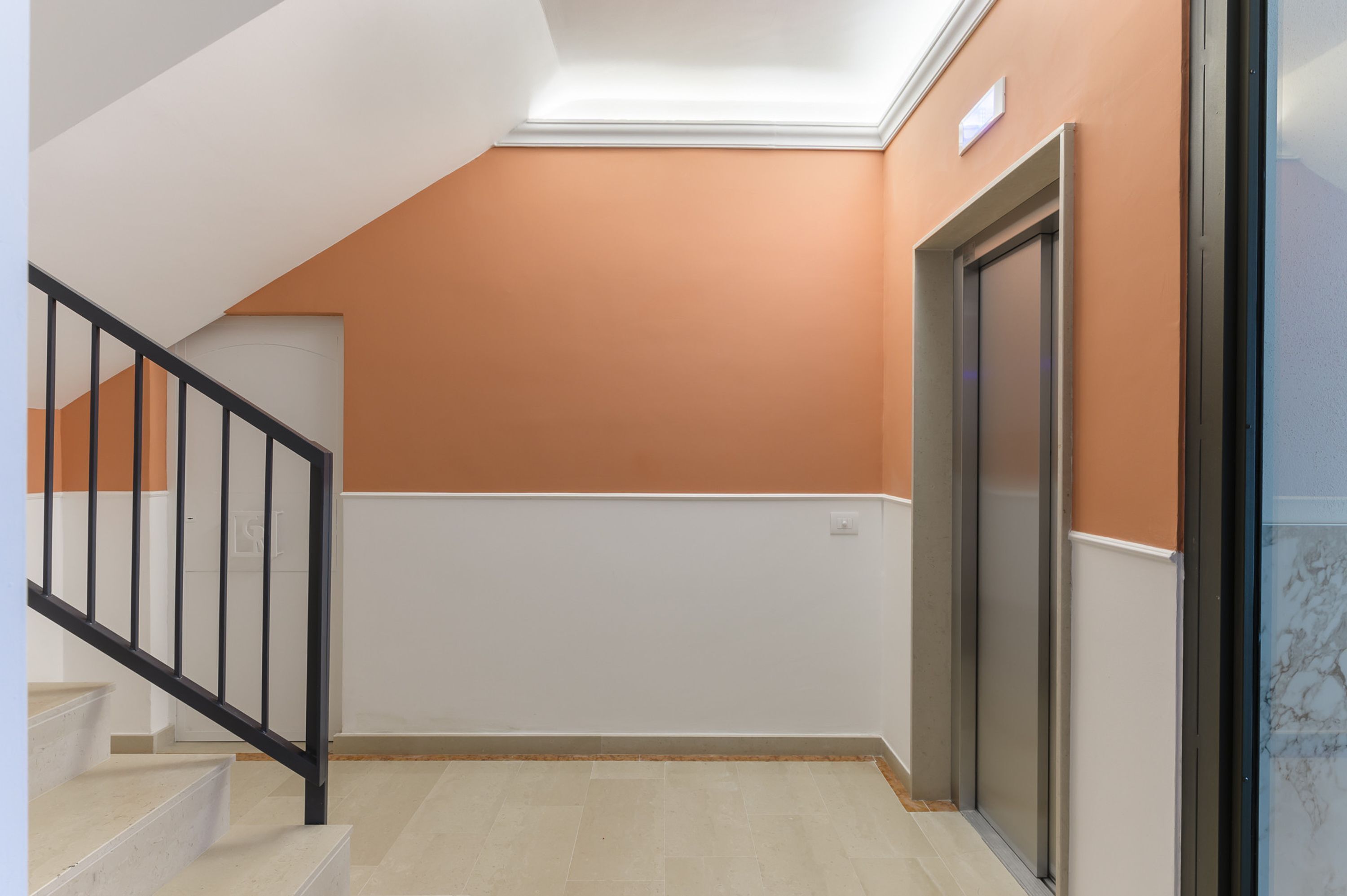 Un escalier avec des rampes noires menant à un palier doté d’un mur blanc et d’un mur orange.