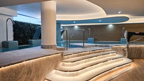Un espace piscine intérieur moderne, avec des marches en marbre menant à un jacuzzi, entouré d’un revêtement de sol en bois et d’un mur recouvert de carreaux bleus.