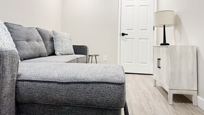 A modern living room with a grey sofa, a small side table, a white door, and a white cabinet. - RiverStone Retreat C (Newburgh)
