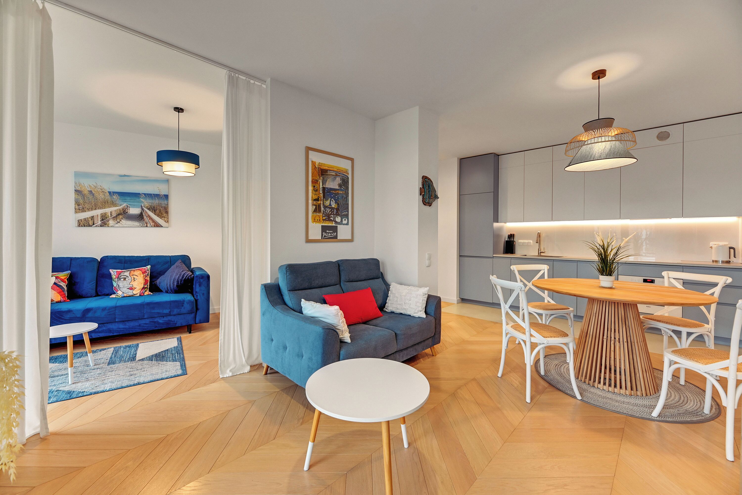 A modern living room with a blue sofa, a grey sofa, a round wooden dining table, and white chairs.