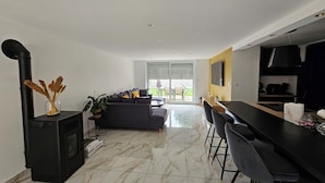 Een moderne woonkamer met een bank, een eethoek met barkrukken, een open haard en een groot raam.
