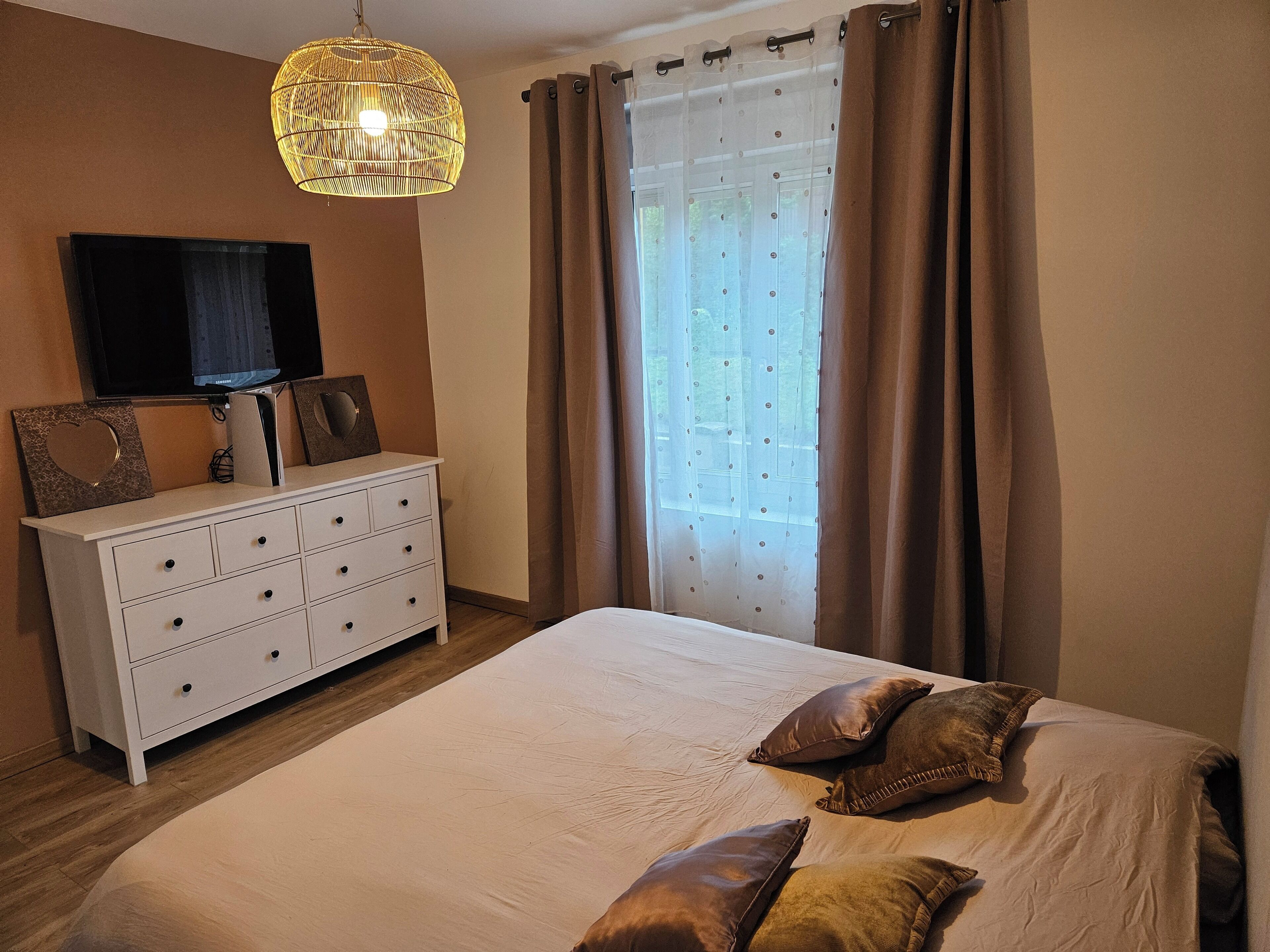 Un dormitorio con una cama, un tocador, un televisor y una ventana con cortinas.