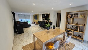 Een ruime woonkamer met een eettafel, stoelen en een bank. Achtergrond is een keuken met een televisie en diverse spullen op de planken.