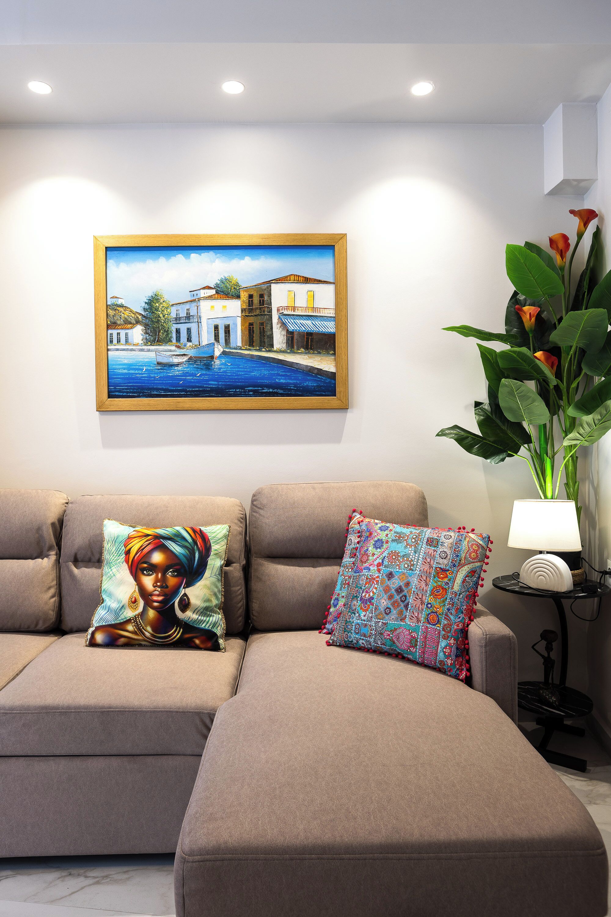 A living room with a beige sofa, two patterned cushions, a framed painting, a potted plant, and a table lamp.
