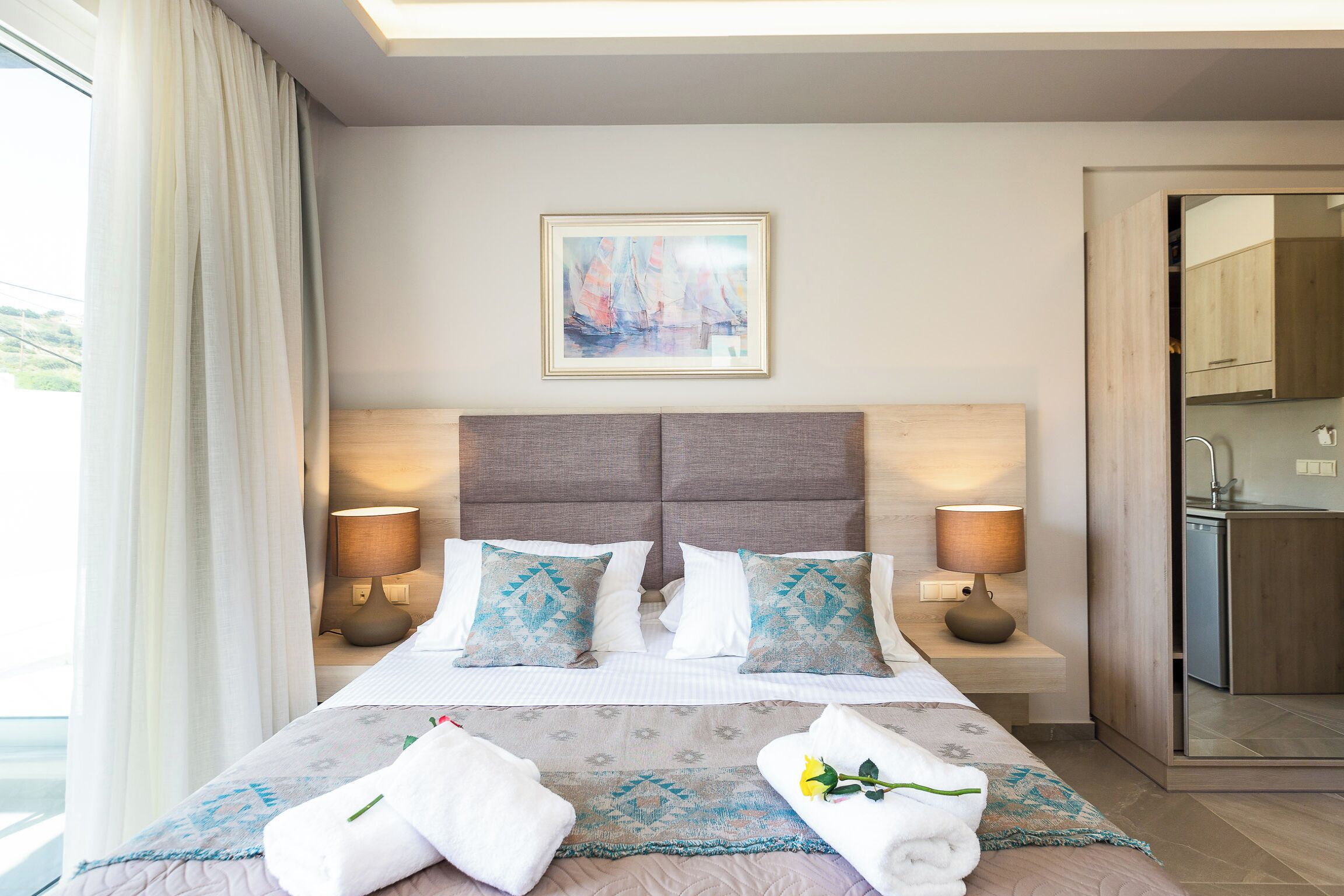 A hotel room with a large bed, two bedside lamps, a framed painting, and a view of the kitchen.