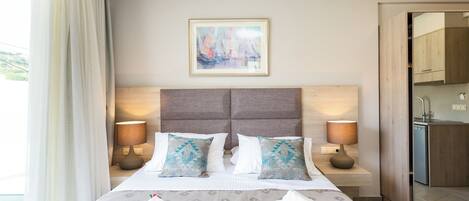 A hotel room with a large bed, two bedside lamps, a framed painting, and a view of the kitchen.